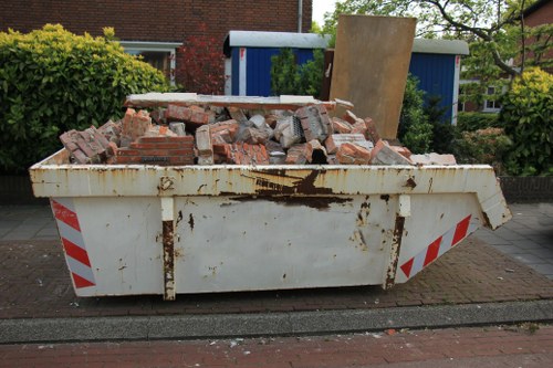 A white, rust-stained skip filled with broken red bricks and rubble, positioned on a paved pavement in a residential area. The debris includes various sizes of bricks, some with rough, weathered textures, and broken mortar joints. The skip is partially overflowing, with some bricks resting at angles, and has red and white reflective stripes on the sides. In the background, there are green bushes, a brick wall, and a blue and white shed with a sloped roof, indicating a typical suburban garden setting. The scene reflects a rubbish removal service in Wandsworth, with the skip ready for disposal or recycling of construction debris, consistent with garden clearance or building refurbishment efforts common in the area.