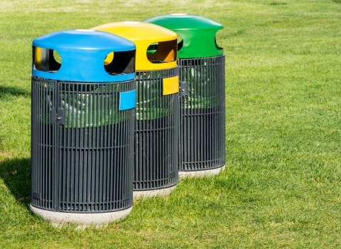 Three cylindrical outdoor trash bins with rounded, brightly coloured lids—blue, yellow, and green—are positioned on a well-maintained grassy area, likely on a pavement or garden within Wandsworth. Each bin features vertical black plastic slats forming the body, with small rectangular labels on the front in matching colours to indicate designated waste types. The bins are placed side by side, aligned in a row, with their foot pedals at the base for easy access. The environment is clearly outdoors, with lush green grass surrounding the bins and no other objects or structures visible in the background. The lighting appears natural, suggesting a clear day. This scene reflects a typical communal outdoor waste disposal point, possibly serving a residential or public space in Wandsworth, with the bins maintained by Garden Clearance Wandsworth, specialists in rubbish management and waste removal services in the local area.