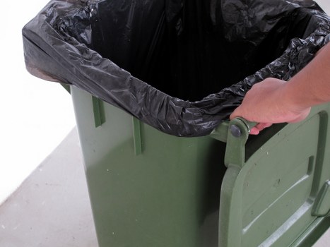 A close-up view of a green wheeled rubbish bin with a black plastic liner inside, positioned on a paved surface, likely in a garden or outdoor area in Wandsworth. A person's hand is gripping the grey handle, indicating the bin is being prepared for waste collection or clearance. The bin's surface shows a matte finish, and the black liner appears to be made of thin, flexible plastic, partially draped over the rim. The environment appears to be an outdoor space, possibly a driveway or garden path, consistent with garden clearance activities undertaken by local waste removal services such as Garden Clearance Wandsworth. The scene reflects a typical scene of rubbish disposal or garden waste collection, with the focus on proper containment of waste materials for removal, supporting the context of professional rubbish removal in the Wandsworth area.