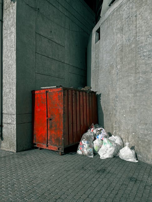 A rusty red metal waste container positioned against a dark green building wall and a light grey concrete wall on a paved city sidewalk. Next to the container, several clear plastic bags filled with m