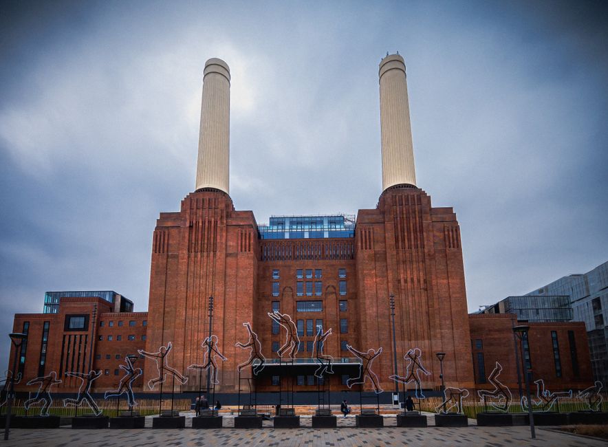 The image shows a close-up view of a large industrial building with a brick and concrete exterior, featuring tall, narrow windows and vertical architectural details. A prominent white chimney or smoke