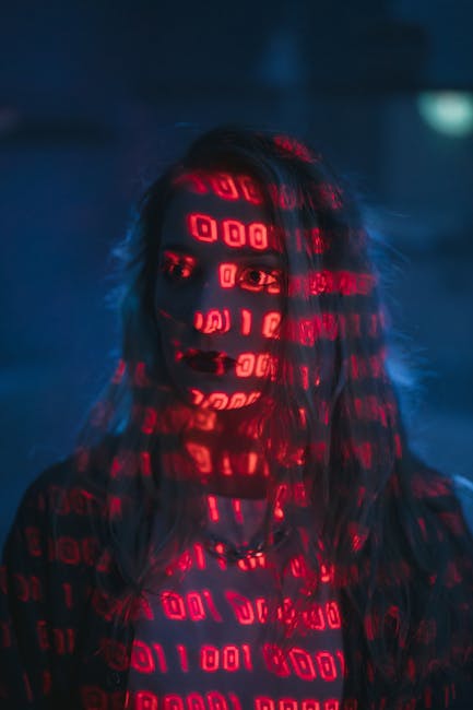 A person with long, dark hair standing in a dark environment, their face obscured by a digital projection of bright red binary code on their face and clothing, creating an abstract, technological visu